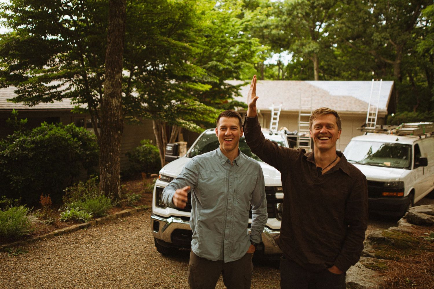 Two smiling men giving a high five outdoors near trucks and a house surrounded by trees.