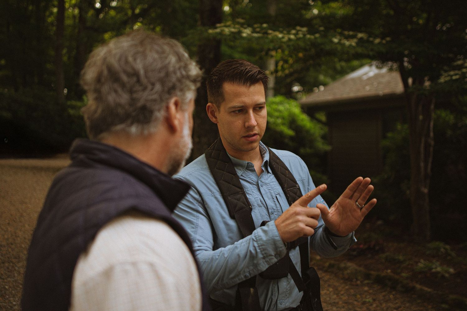 Two men having an outdoor conversation near a wooded area, one gesturing with hands while wearing a camera strap.