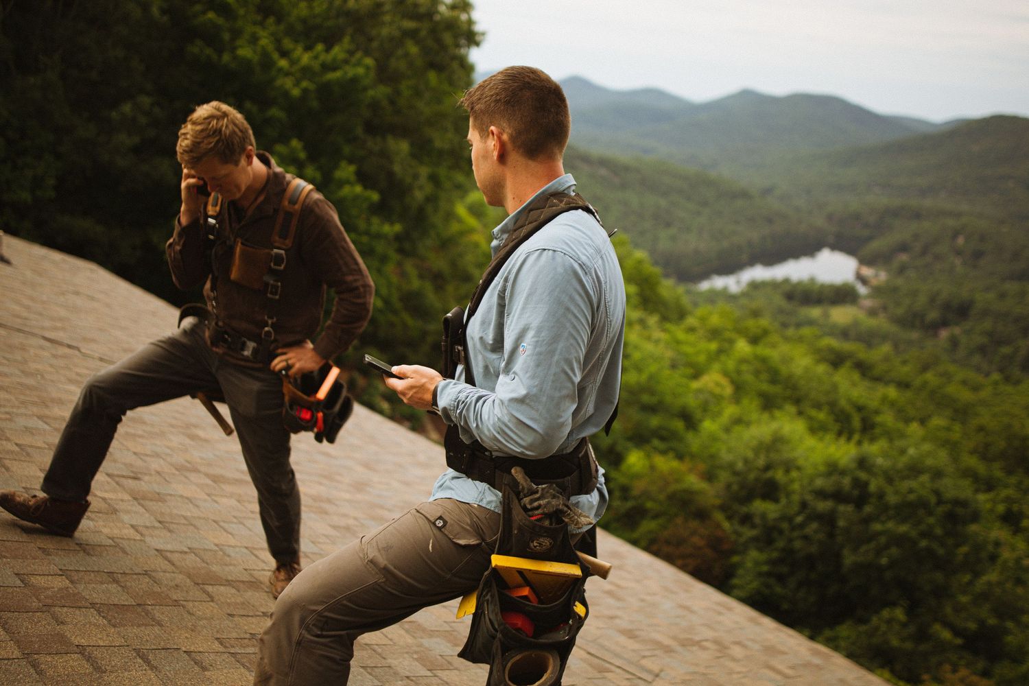 Two roofers with tool belts working on a sloped roof with scenic mountain and lake views in the background.