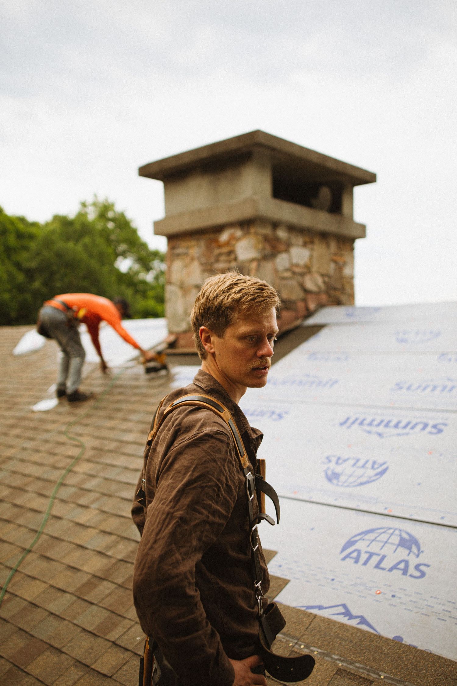 Two roofers installing Atlas Summit underlayment on a residential roof near a stone chimney on an overcast day.