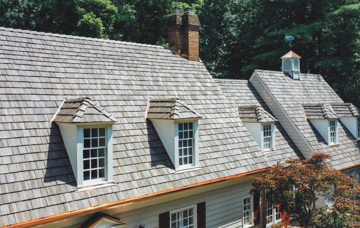 Historic home with gray cedar shake roof featuring dormers, copper weathervane, and brick chimney surrounded by lush trees.