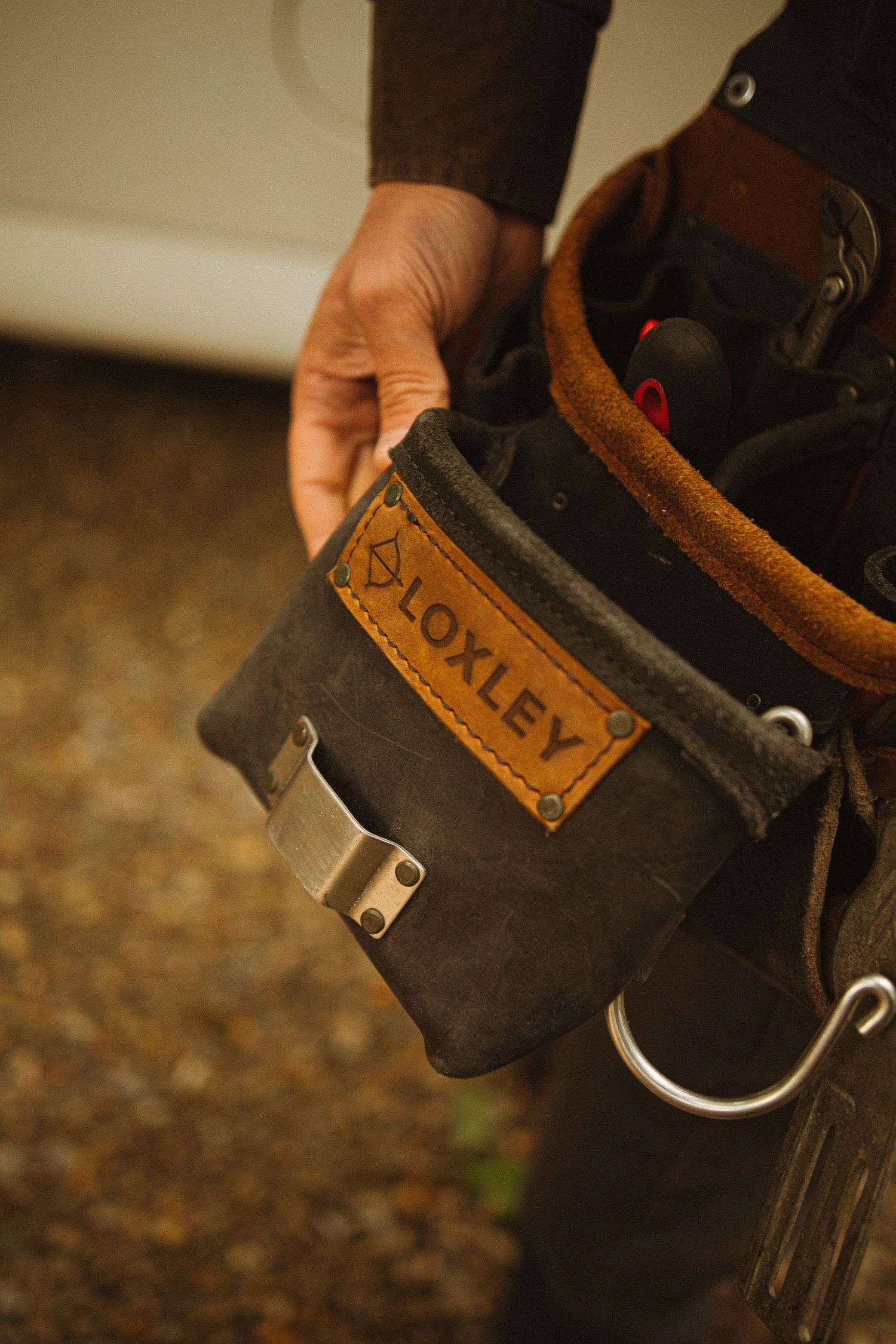 Close-up of a Loxley branded leather tool belt pouch worn by a tradesperson, featuring metal clips and buckles.