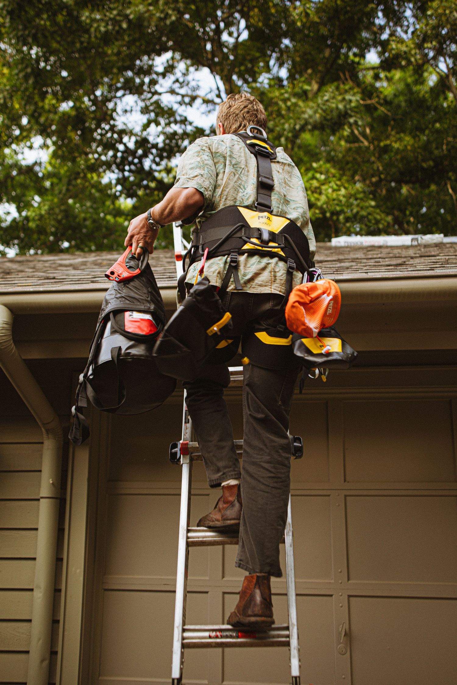 Worker climbing ladder to rooftop wearing safety harness and tool belt for home repair maintenance.