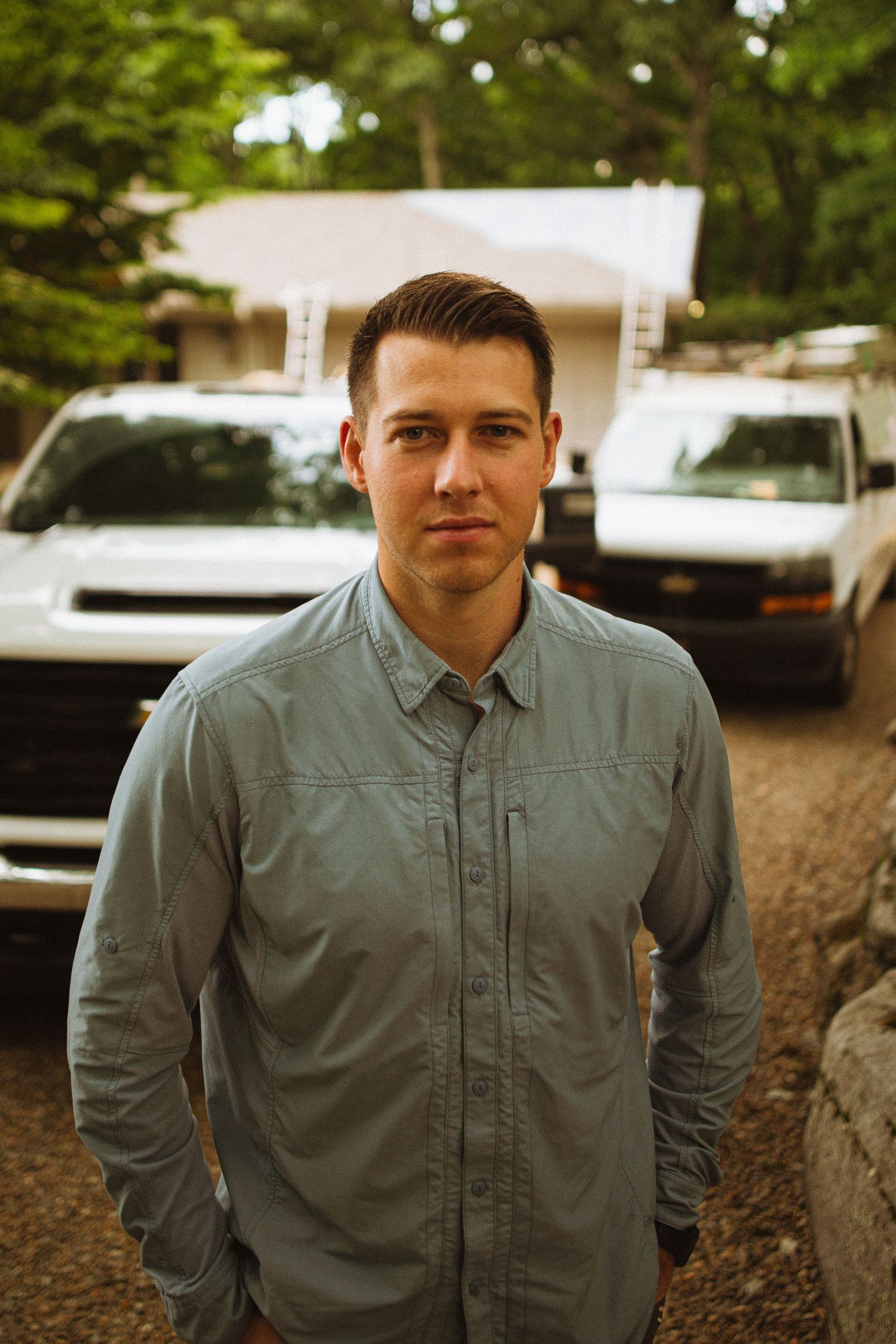 Young man in gray button-up shirt stands confidently outdoors with white trucks and house in background.
