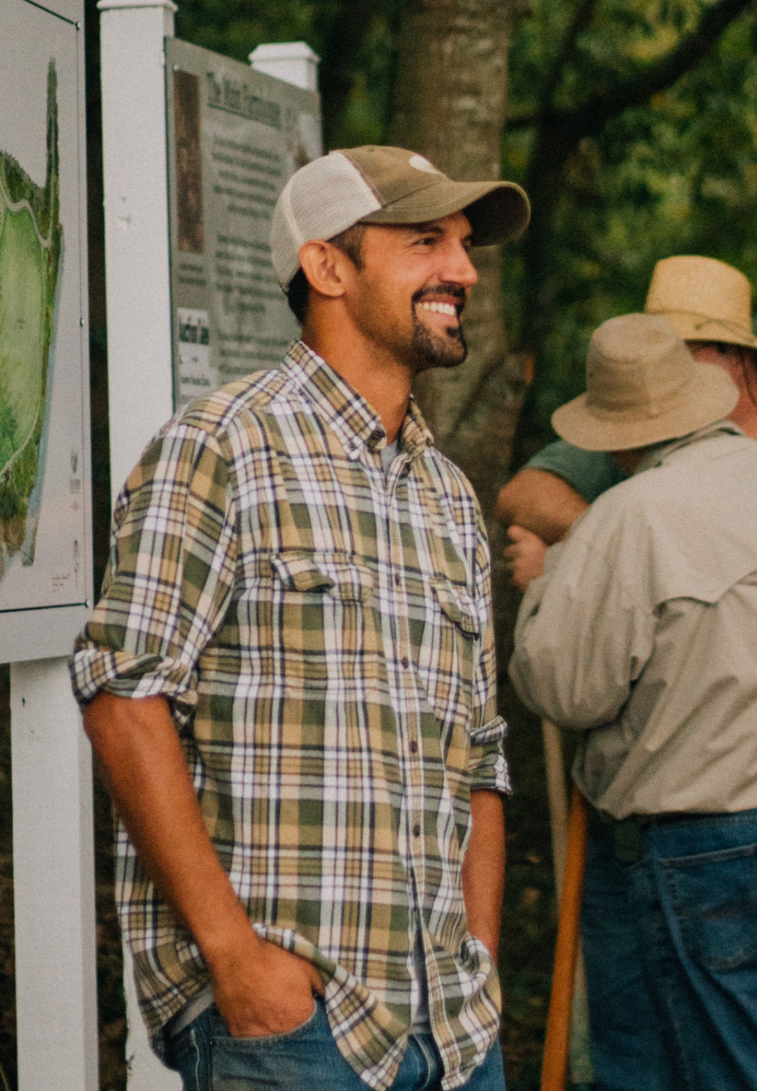 Smiling man in plaid shirt and cap stands outdoors near an informational sign, with people in background.