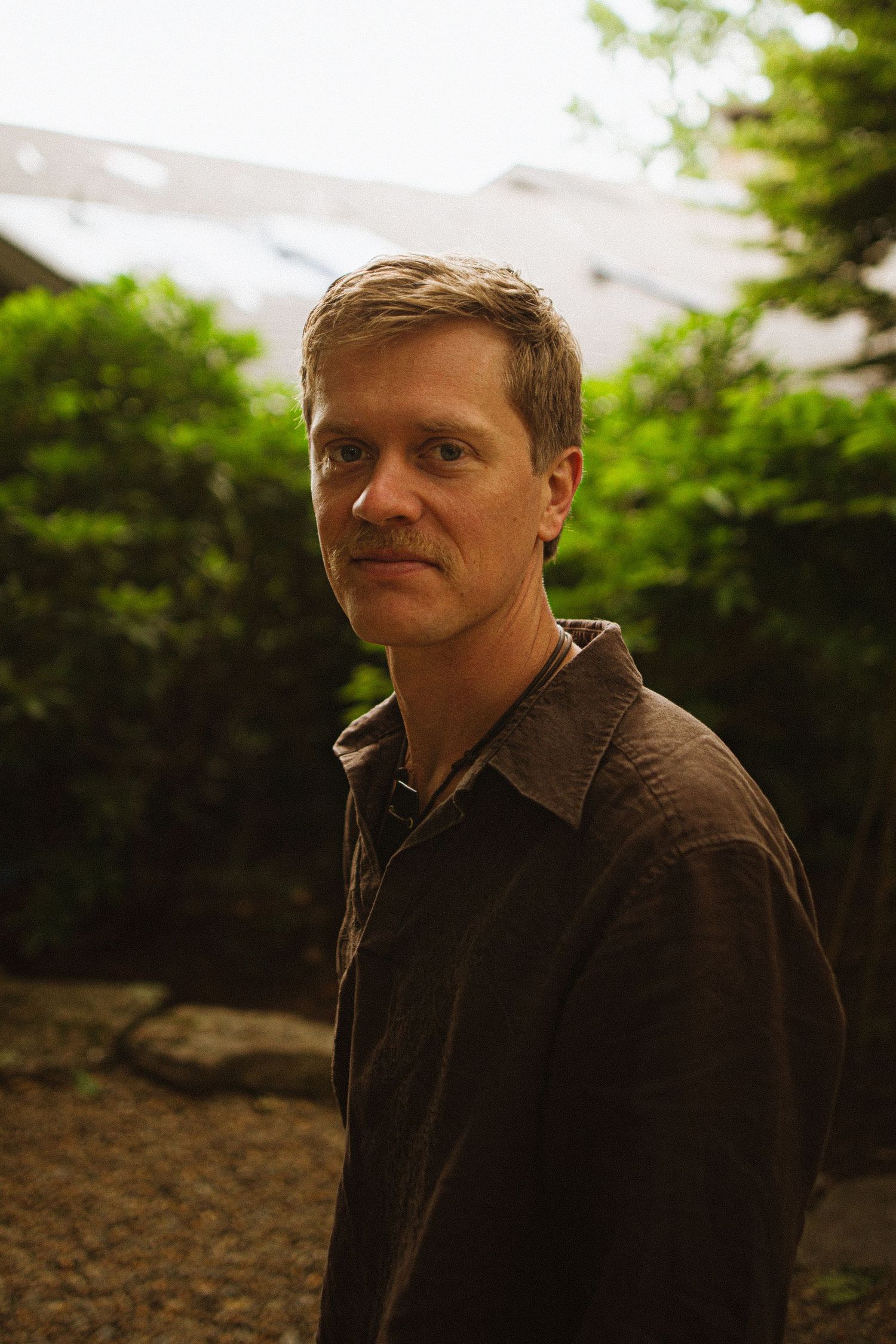 A man in a dark brown jacket stands outdoors among lush green trees, looking over his shoulder in natural light.