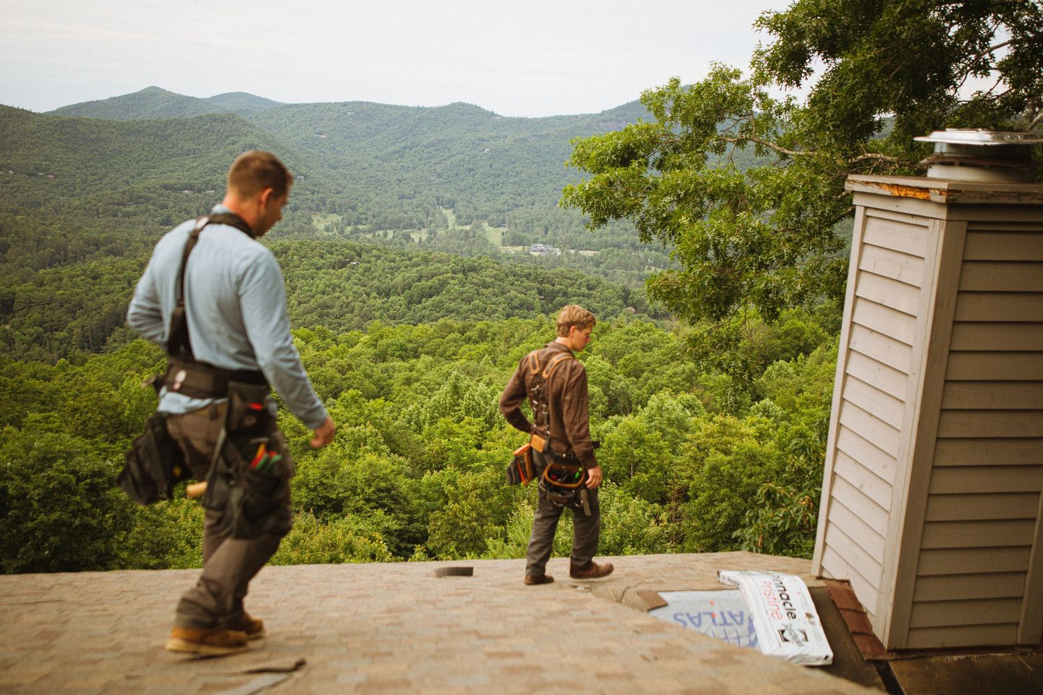 Two workers in tool belts walk on a rooftop with a scenic mountain forest view in the background.