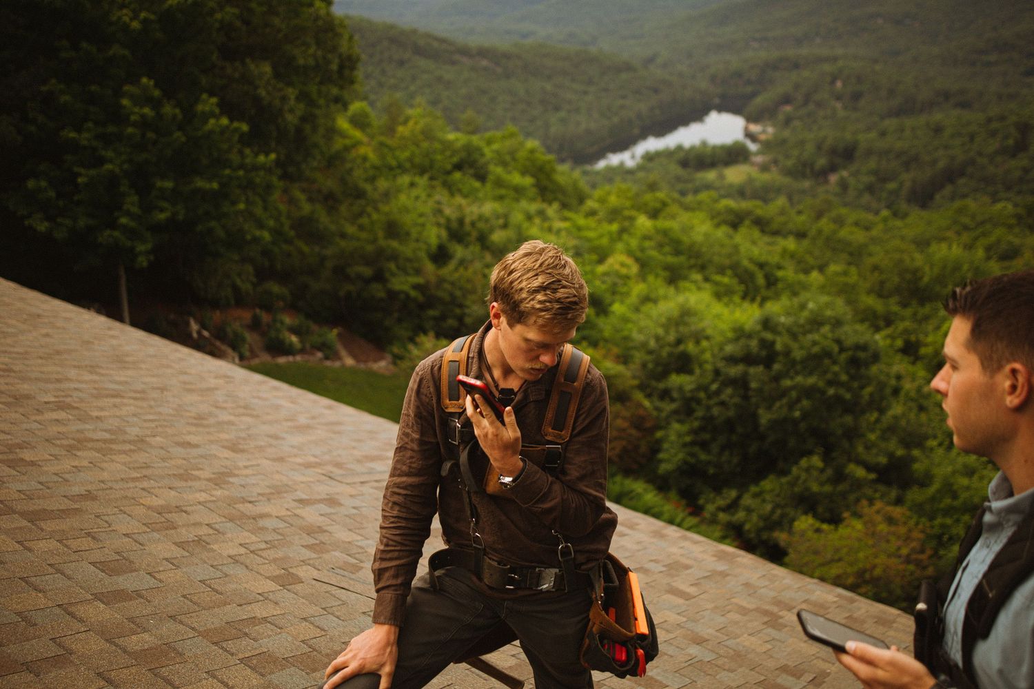 Two men on a rooftop with scenic forest and river views, one holding tools in work gear.
