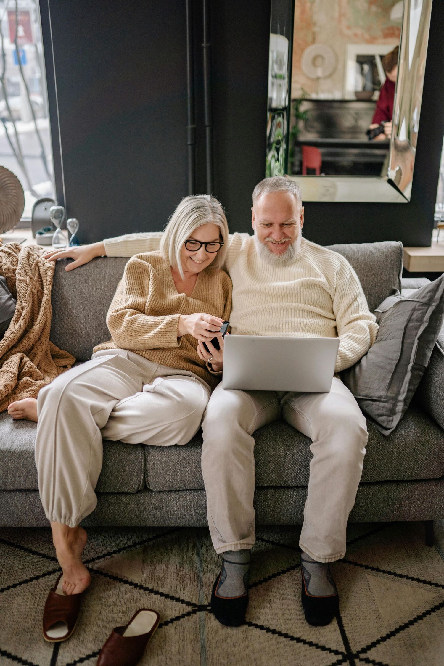 Two people in matching cream sweaters and white pants sit casually on a gray couch while looking at electronic devices.