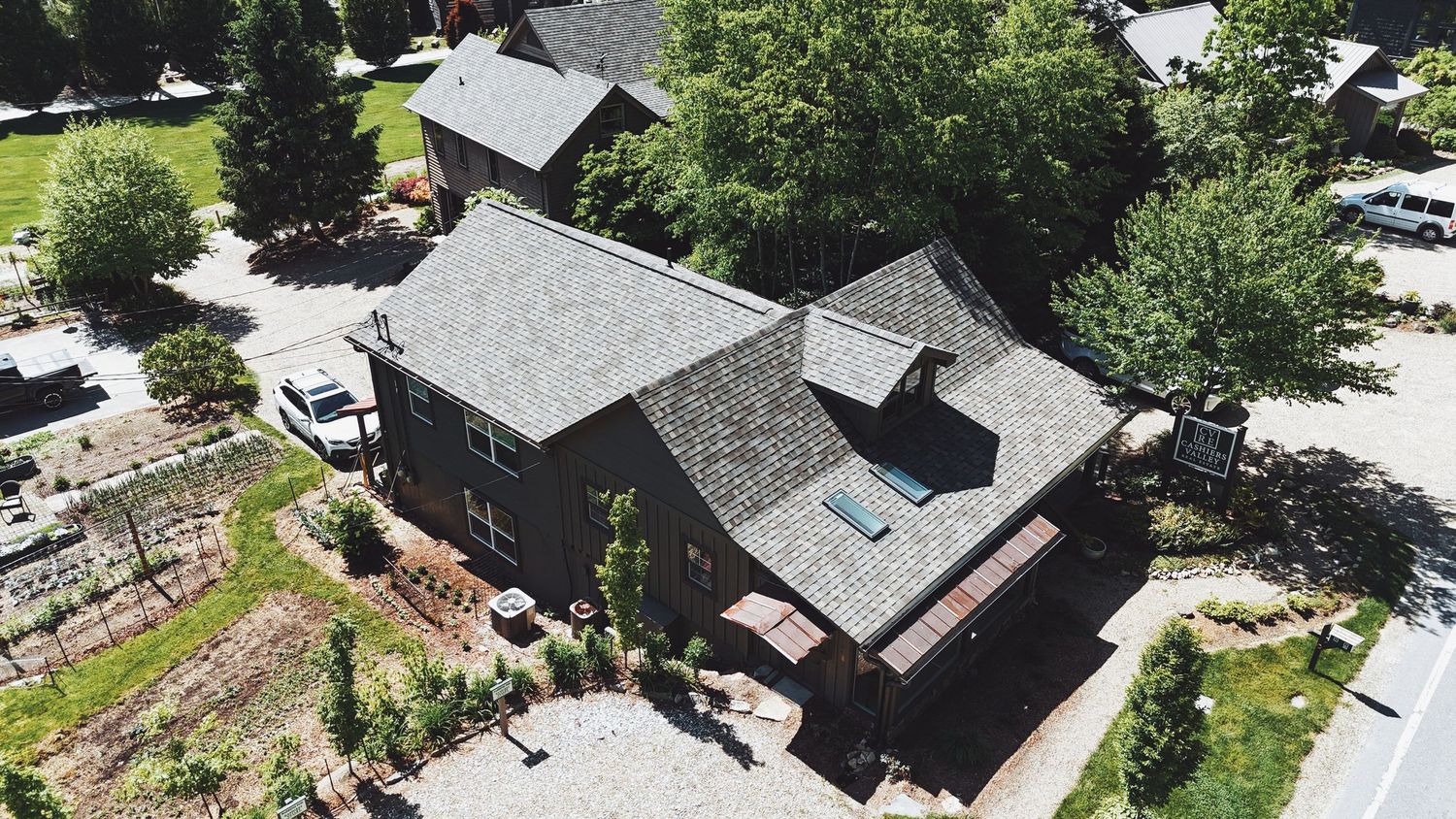Aerial drone view of a dark-shingled residential home surrounded by mature trees and landscaped yard.
