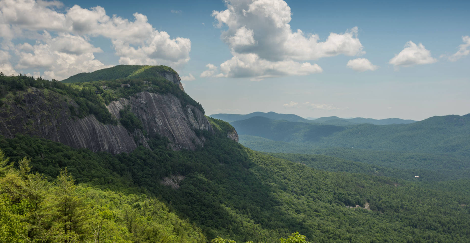 Panoramic mountain landscape with green forests and dramatic rock formations under blue sky.
