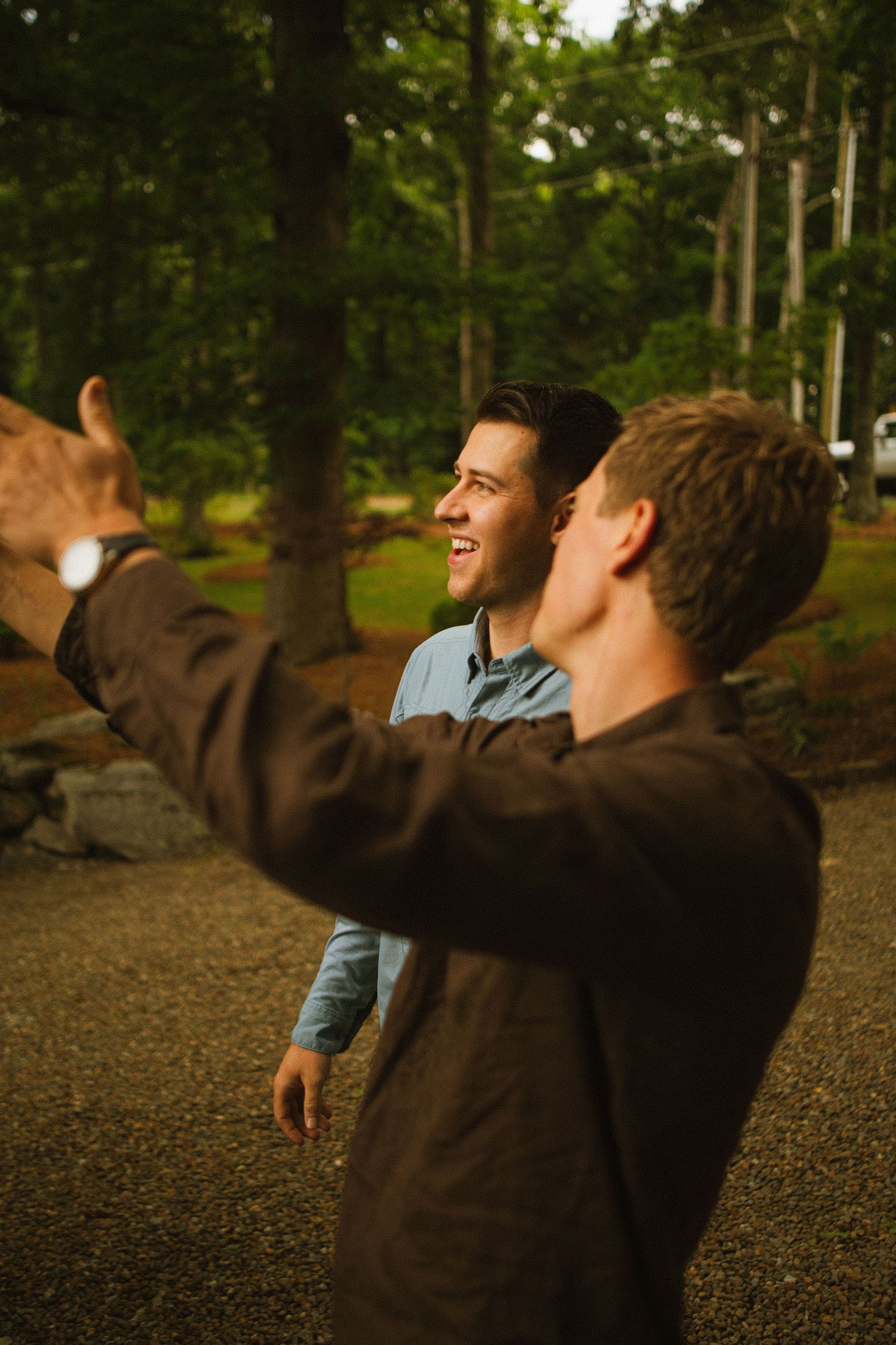 Two young men smiling and pointing outdoors near tall green trees in a forest setting.