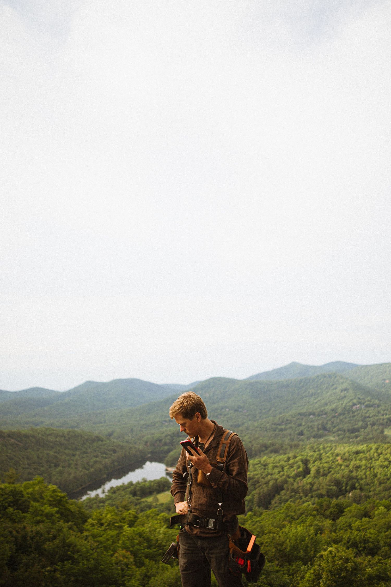 A photographer reviews his camera while standing on a scenic overlook with lush green mountains and a river below.