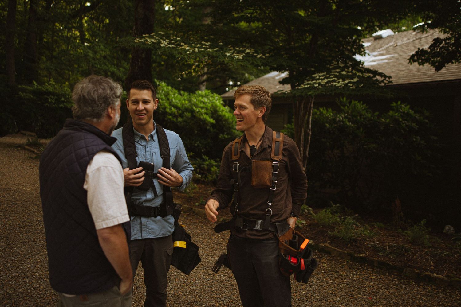 Three men conversing outdoors in a wooded area, two holding camera equipment during an evening photography session.