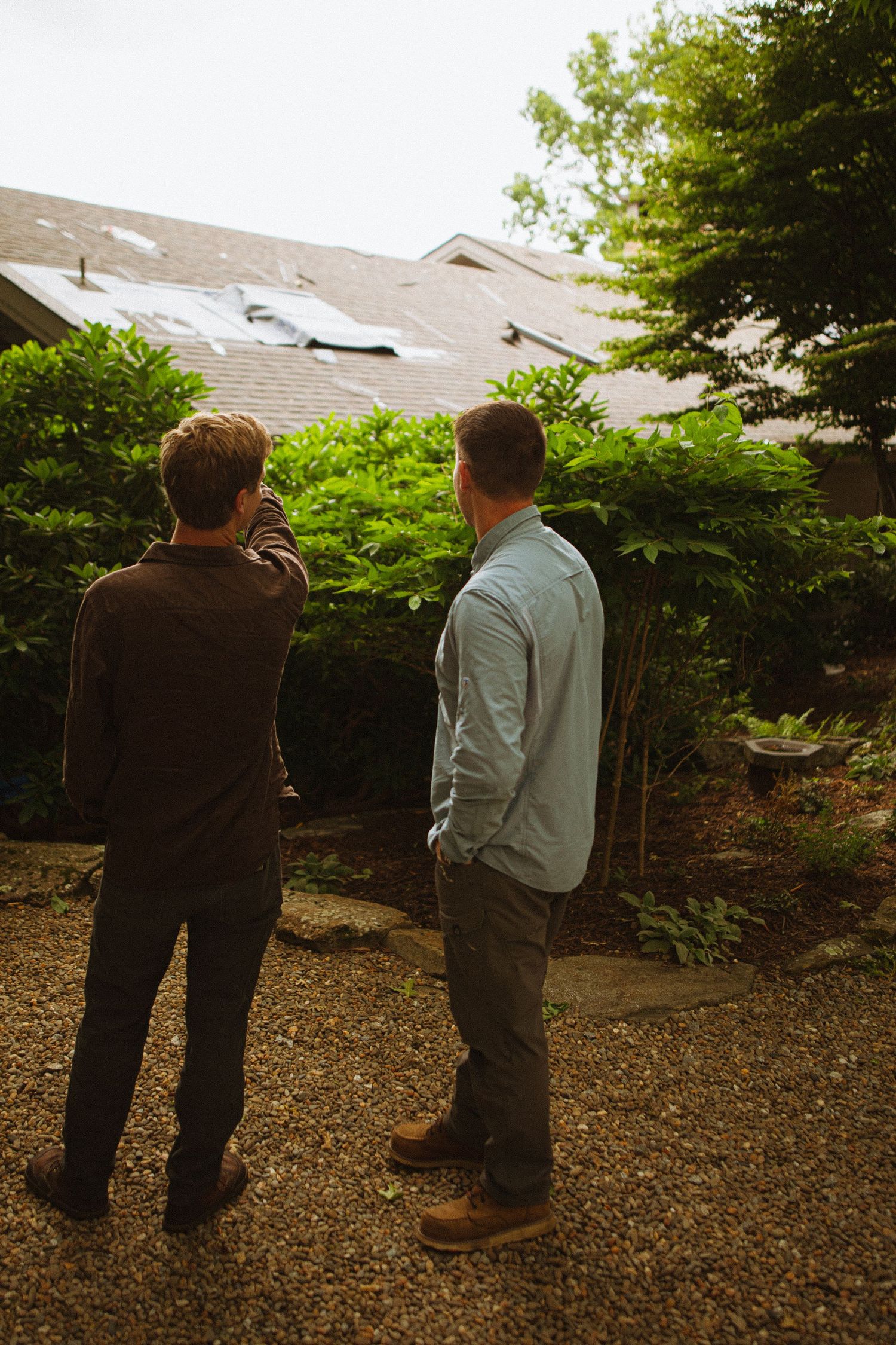Two men standing in a garden examining storm damage on a residential roof surrounded by lush green trees.