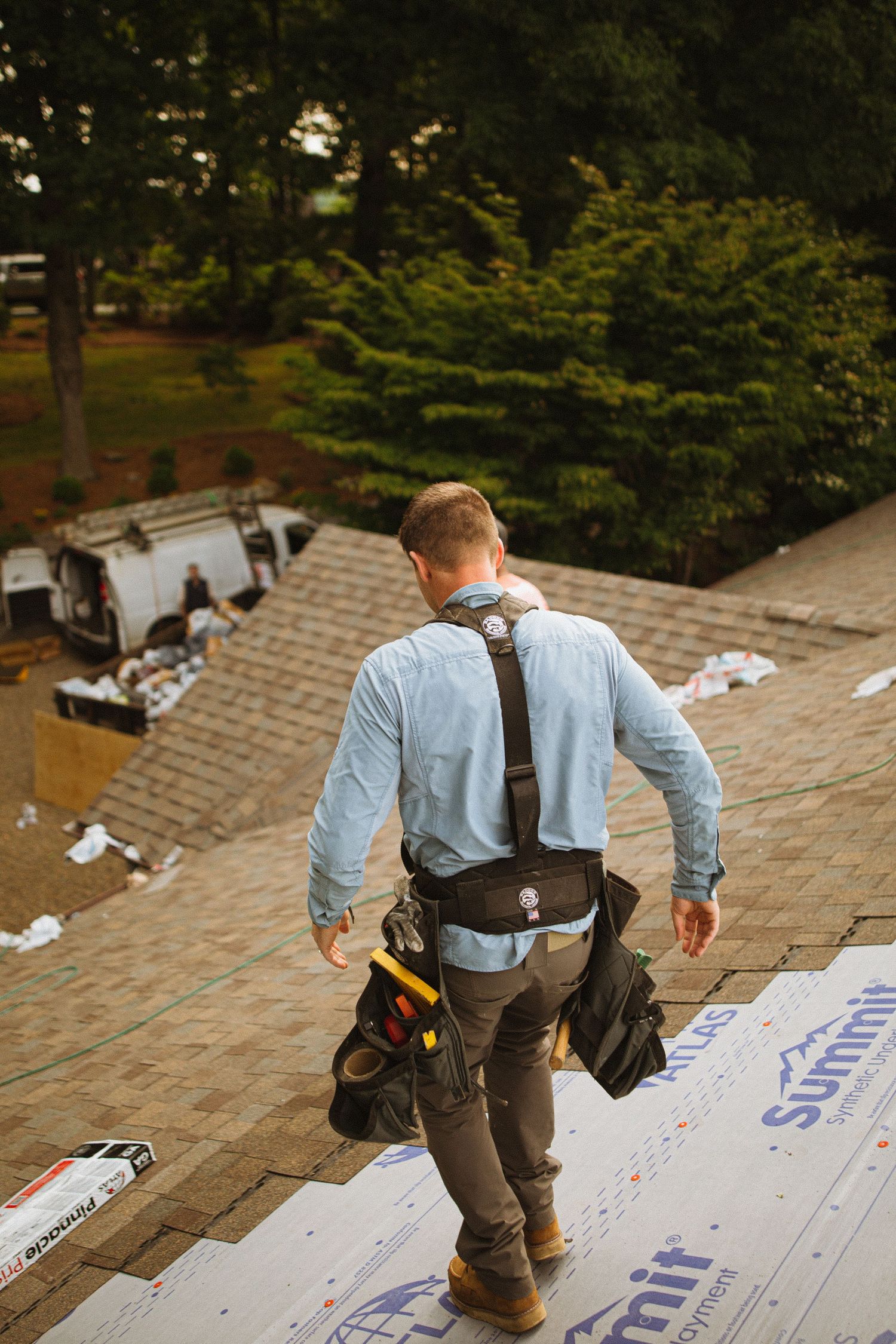 Roofer in tool belt walks across shingled roof during installation, with Summit underlayment visible and trees in background.