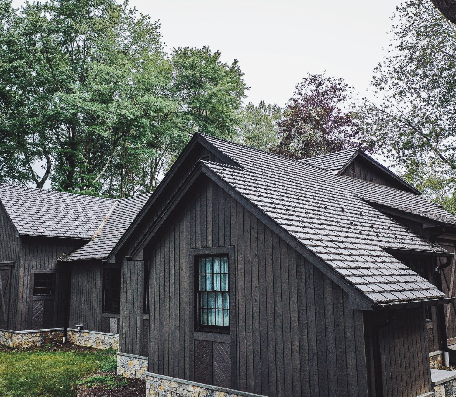 Dark wood cabin with gray shingle roof surrounded by lush green trees, rustic stone wall in foreground.