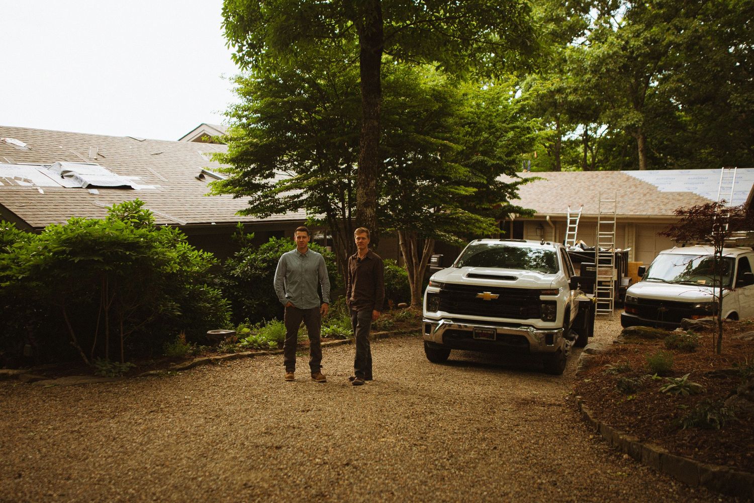 Two contractors stand in a gravel driveway beside a white Chevrolet truck near a house with roofing work underway.