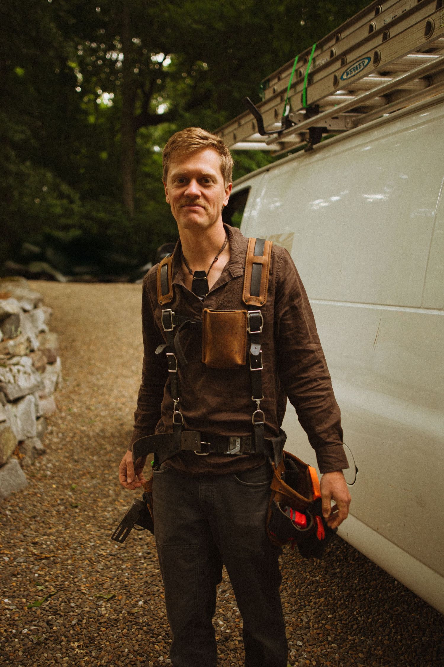 A tradesman wearing leather tool harness stands confidently by a white work van on a gravel driveway.
