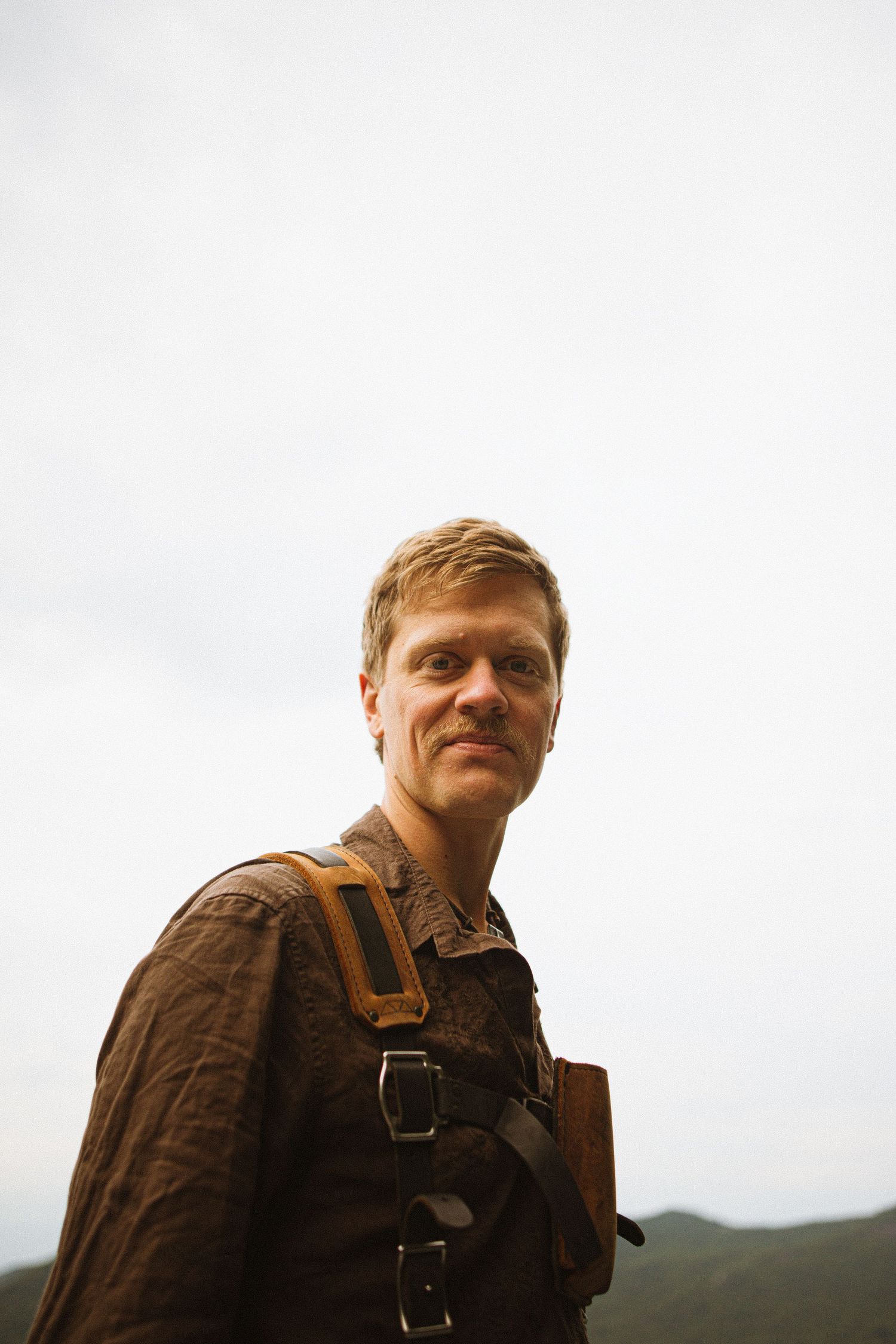 A blond man wearing a brown jacket with leather straps smiles outdoors against a cloudy sky and green hills.