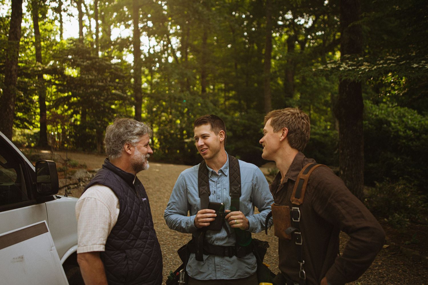 Three men conversing outdoors in a sunlit forest near a vehicle, enjoying nature together.