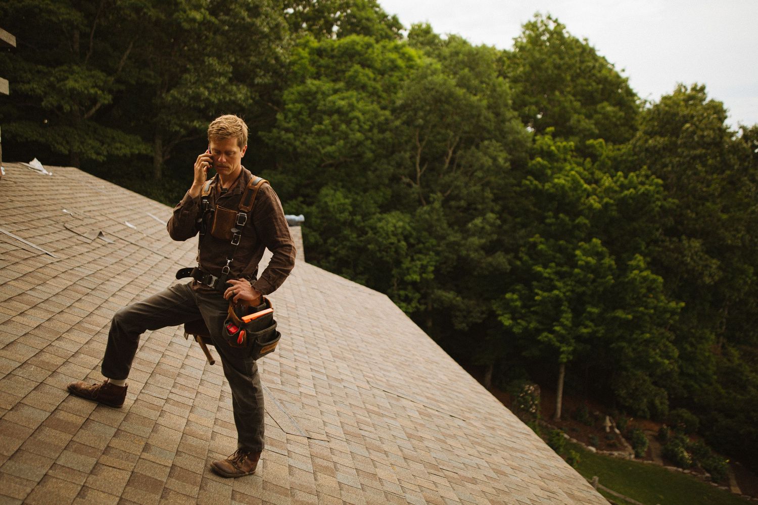 A roofer stands on a shingled roof talking on his phone while holding tools, with lush green trees in the background.