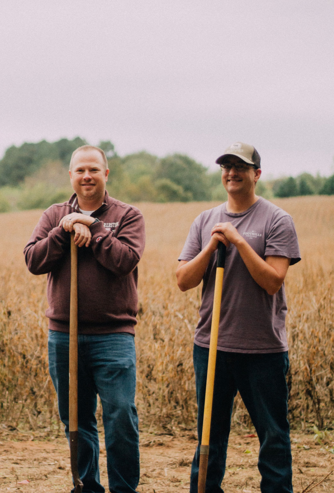 Two farmers smiling in a field holding shovels during harvest season.