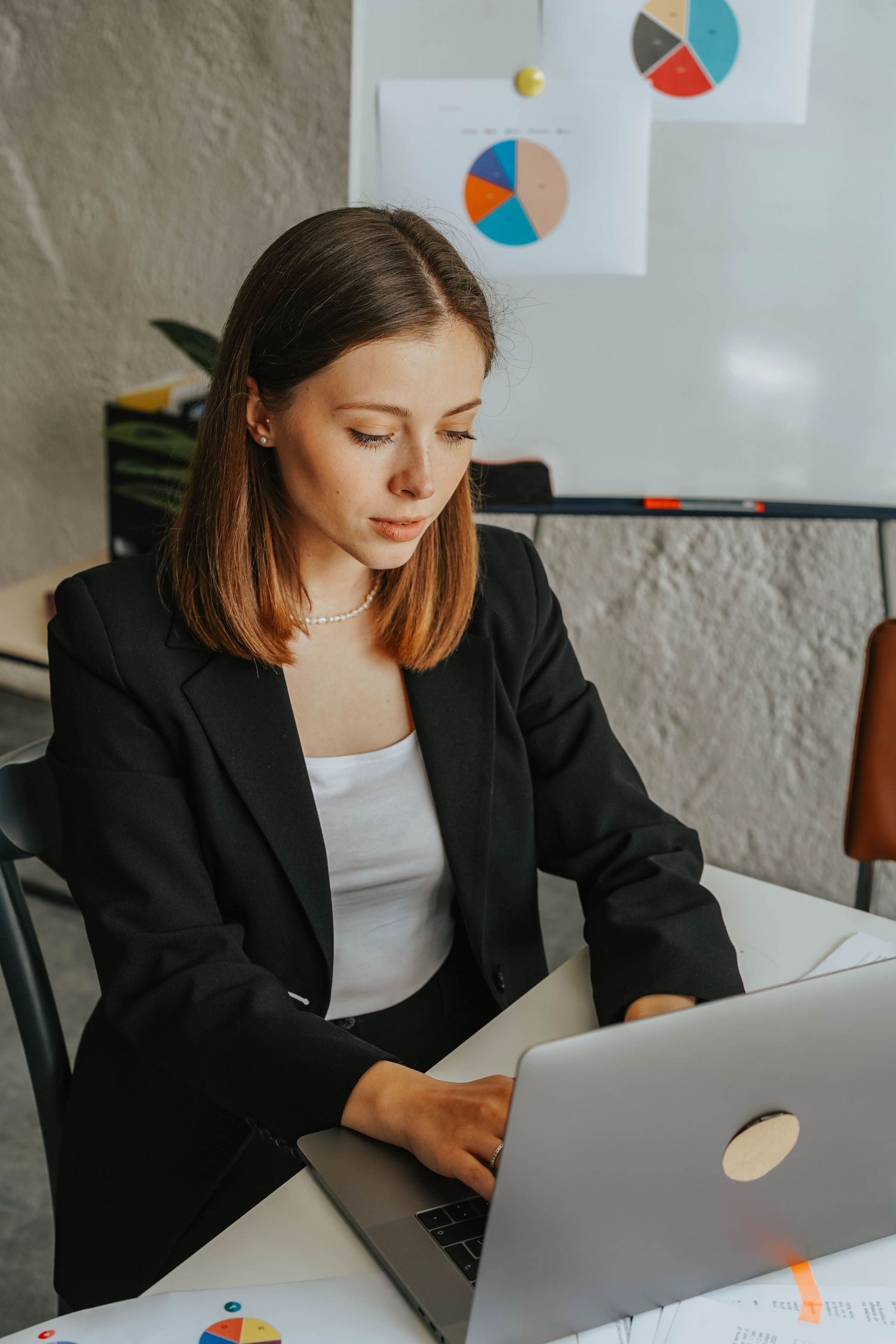 Young businesswoman in black blazer working on laptop at office desk with colorful pie charts on whiteboard behind her.