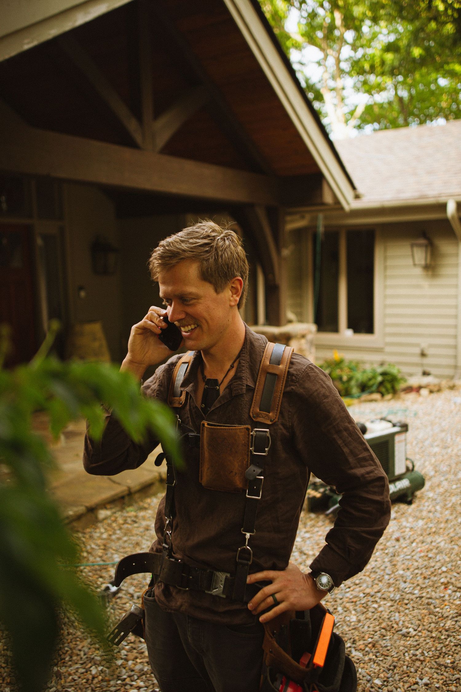 Smiling male contractor in tool belt and suspenders talking on smartphone outside a residential home.
