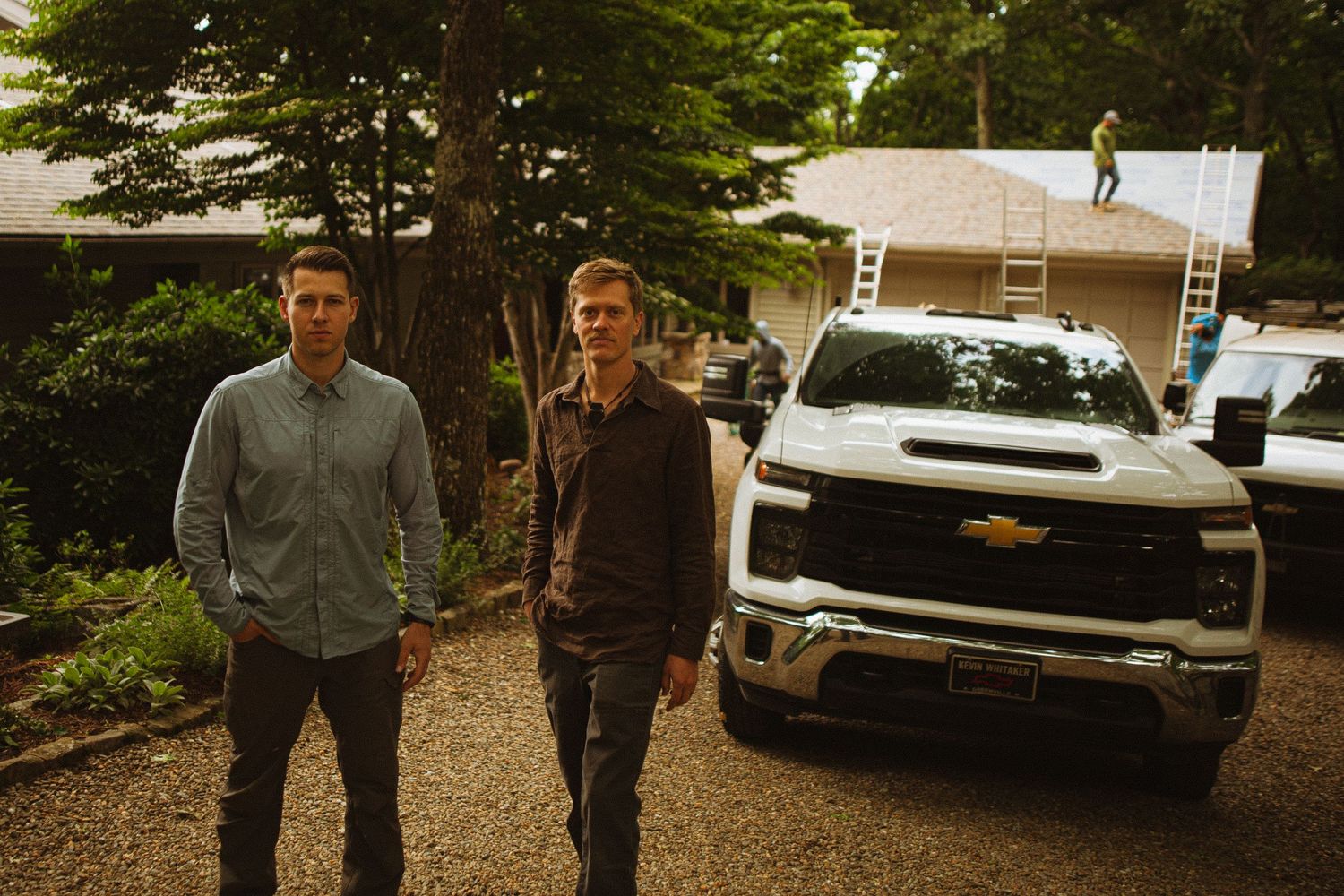 Two men stand beside a white Chevrolet truck on a gravel driveway near a house under roof construction.