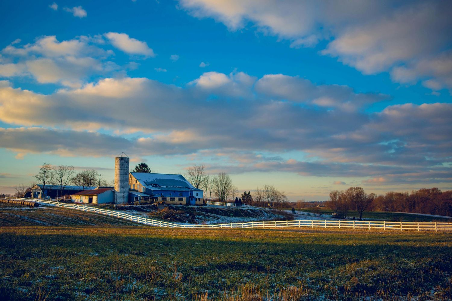 A panoramic view of a rural farm landscape with white barns and silos against a dramatic cloudy blue sky at sunset.