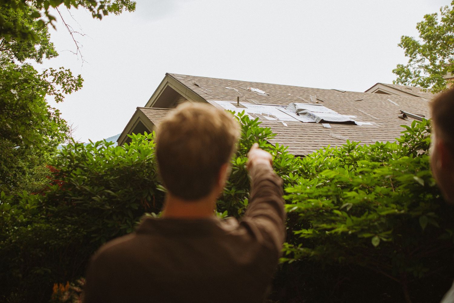Two people pointing at storm damage on a residential roof surrounded by green trees.