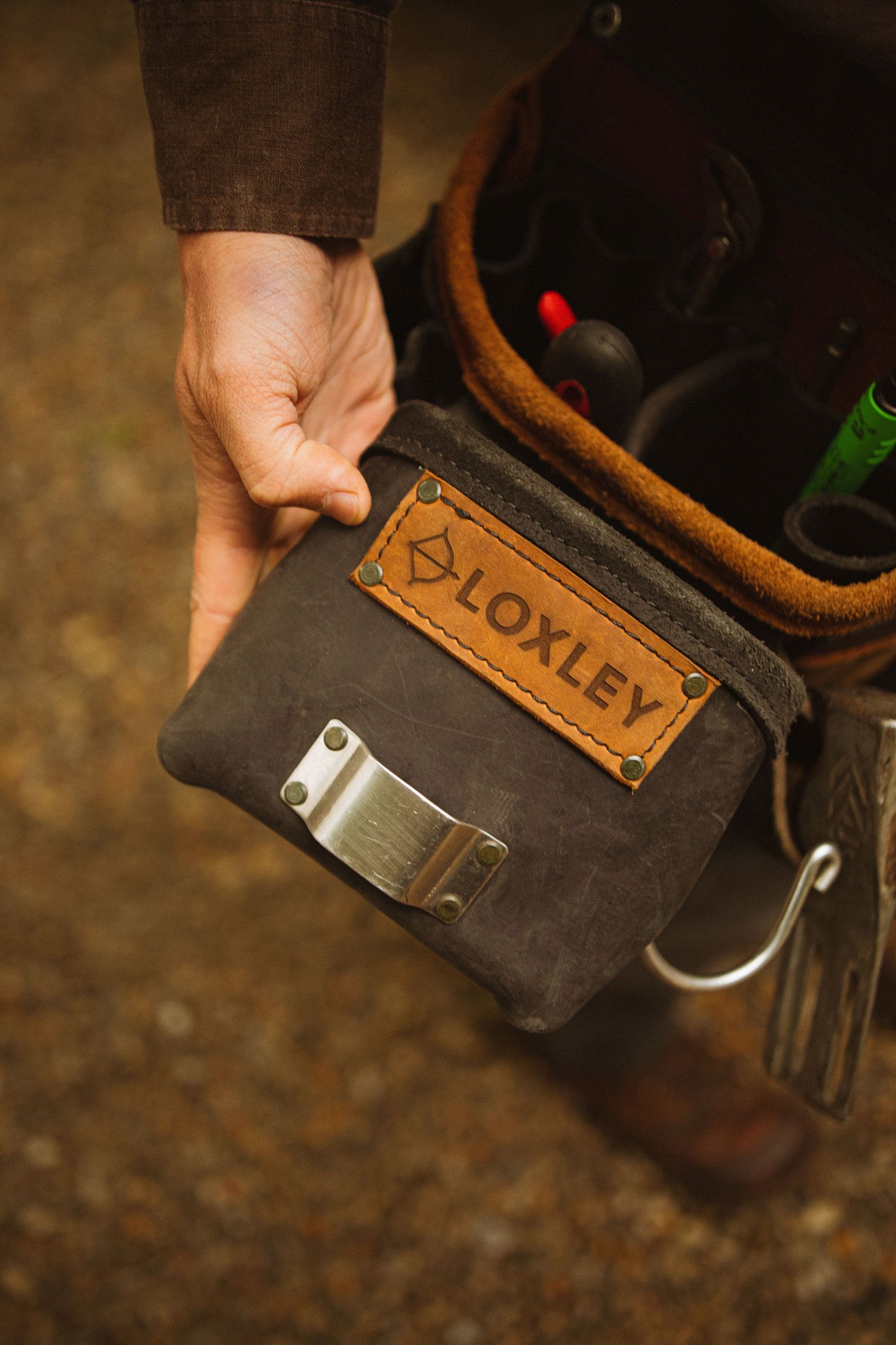 Hand holding a Loxley branded waxed canvas belt pouch with metal clip, next to a leather tool bag.