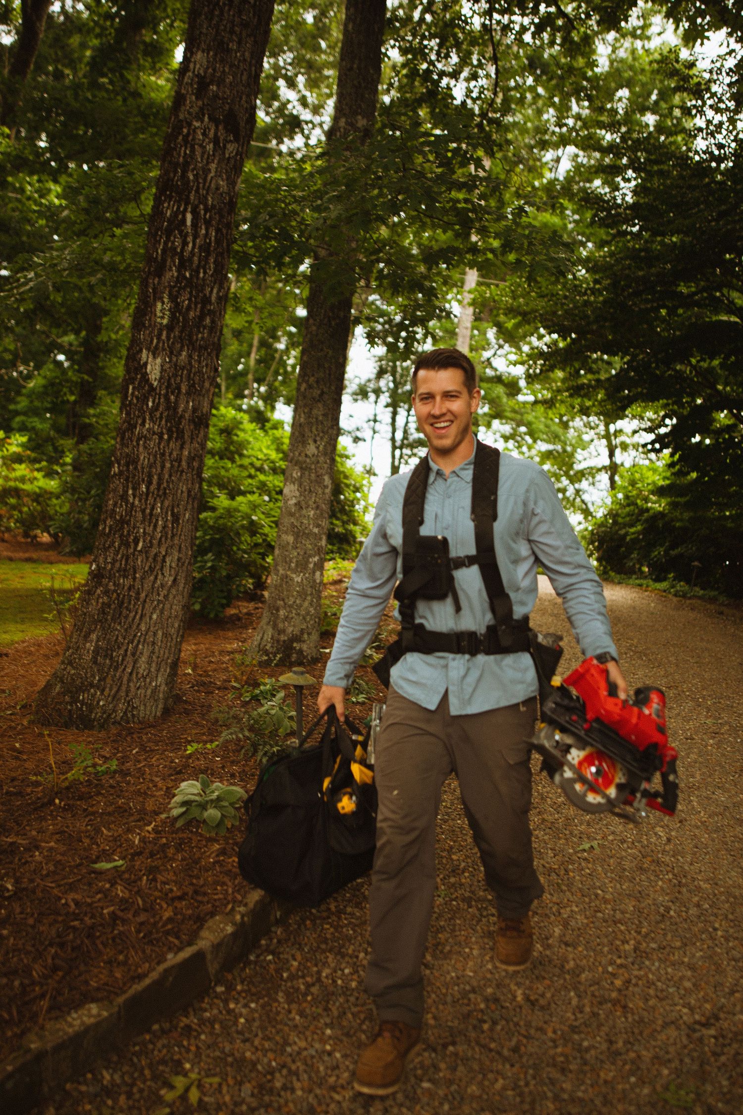 Smiling man carrying camera gear and bags while walking on a forest path surrounded by tall trees.