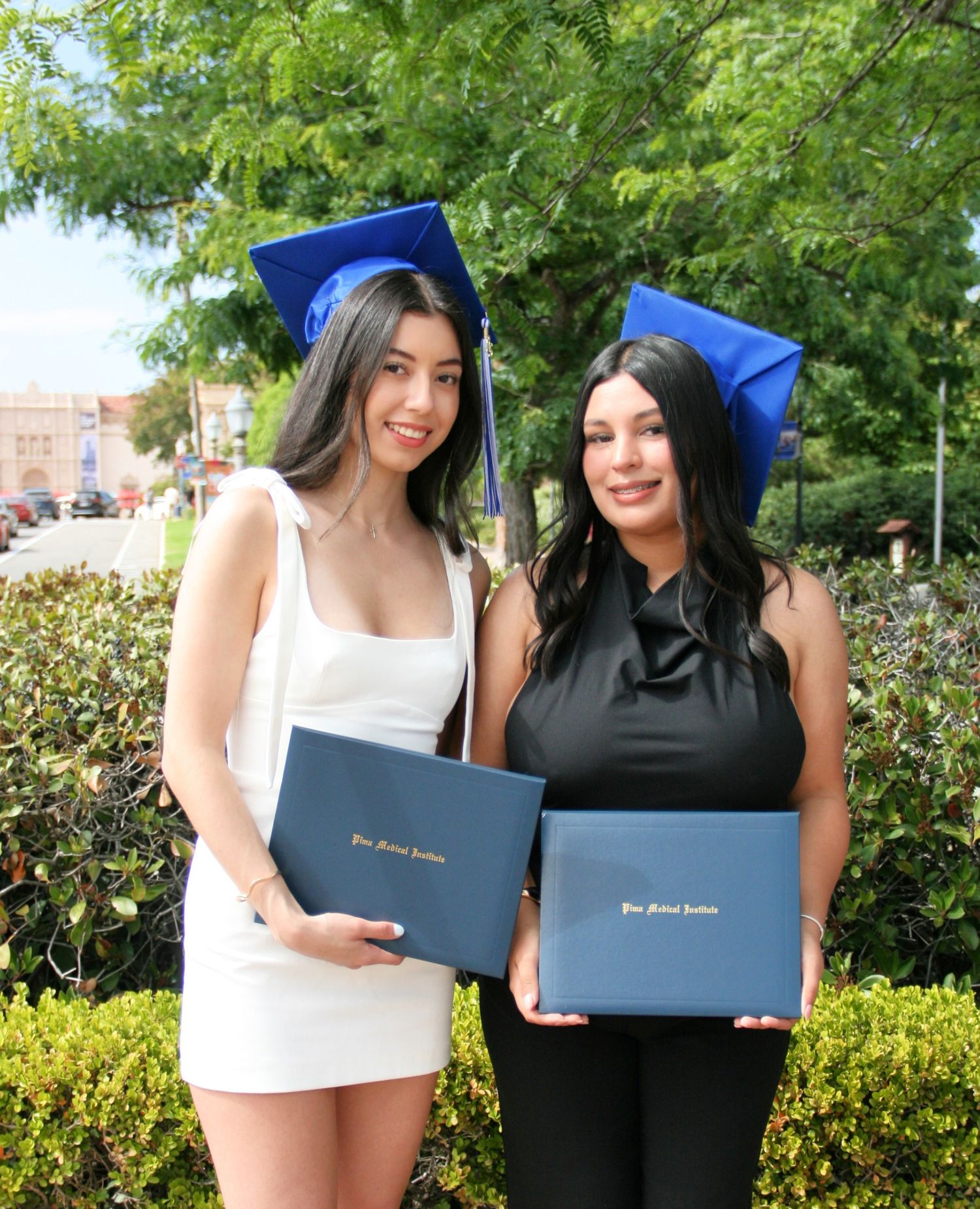 Two graduates in blue caps holding diplomas, smiling outdoors | Nuvue Visuals graduation photography in San Diego, Speckles organ pavillion