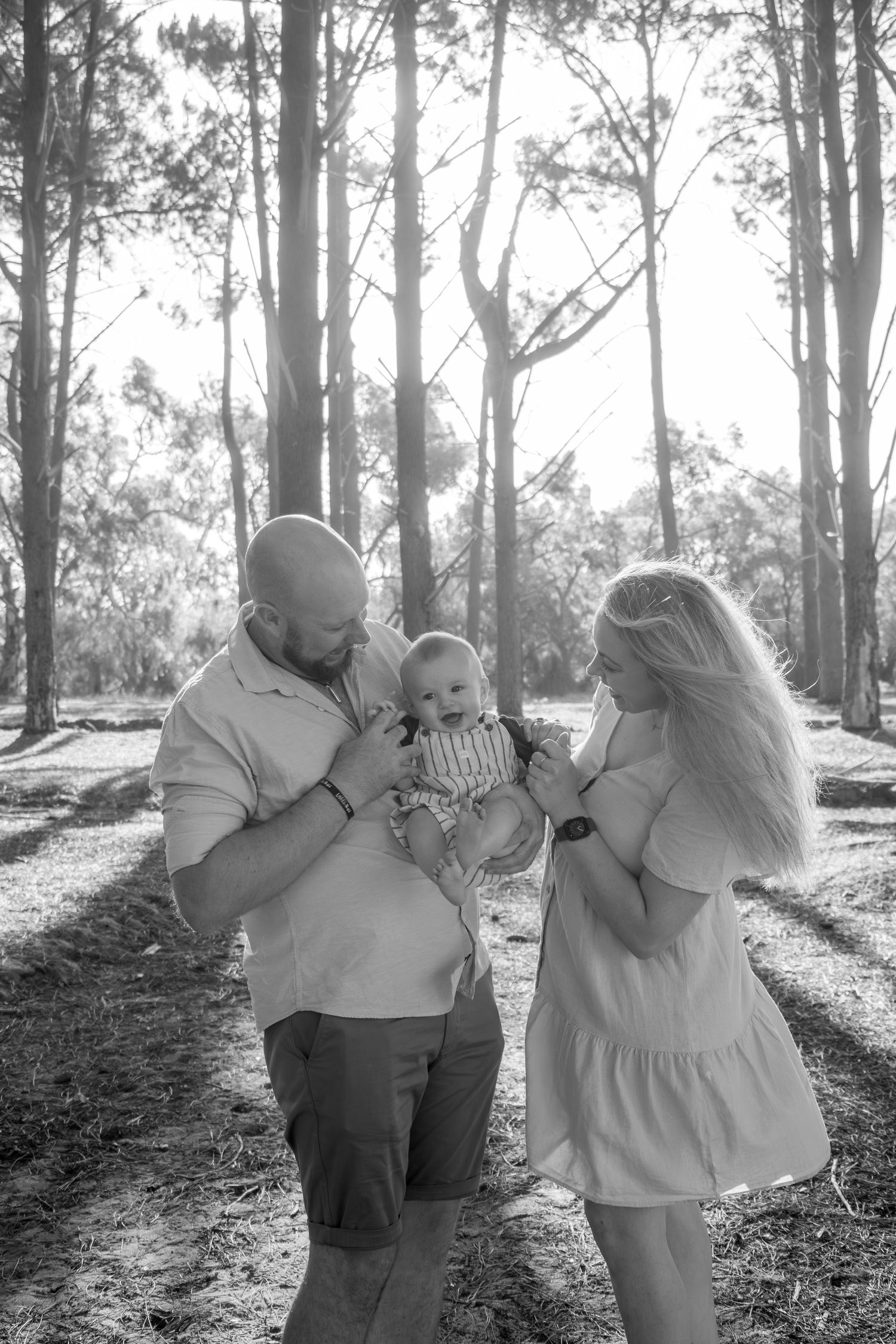 Mum, dad and baby laughing in the pines at sunset during a candid, natural family photography session.
