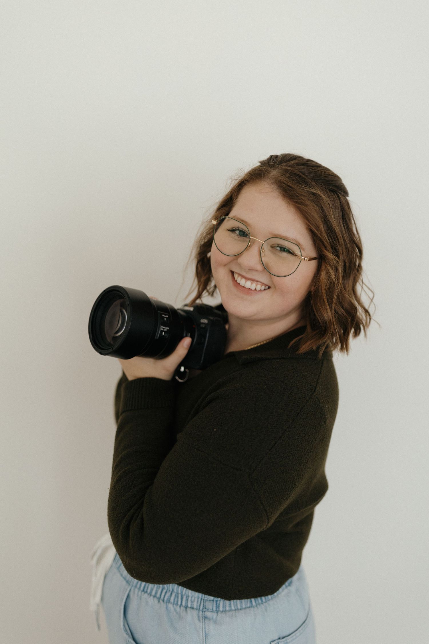 A photographer in a black sweater and jeans holds a professional camera while posing against a white wall.