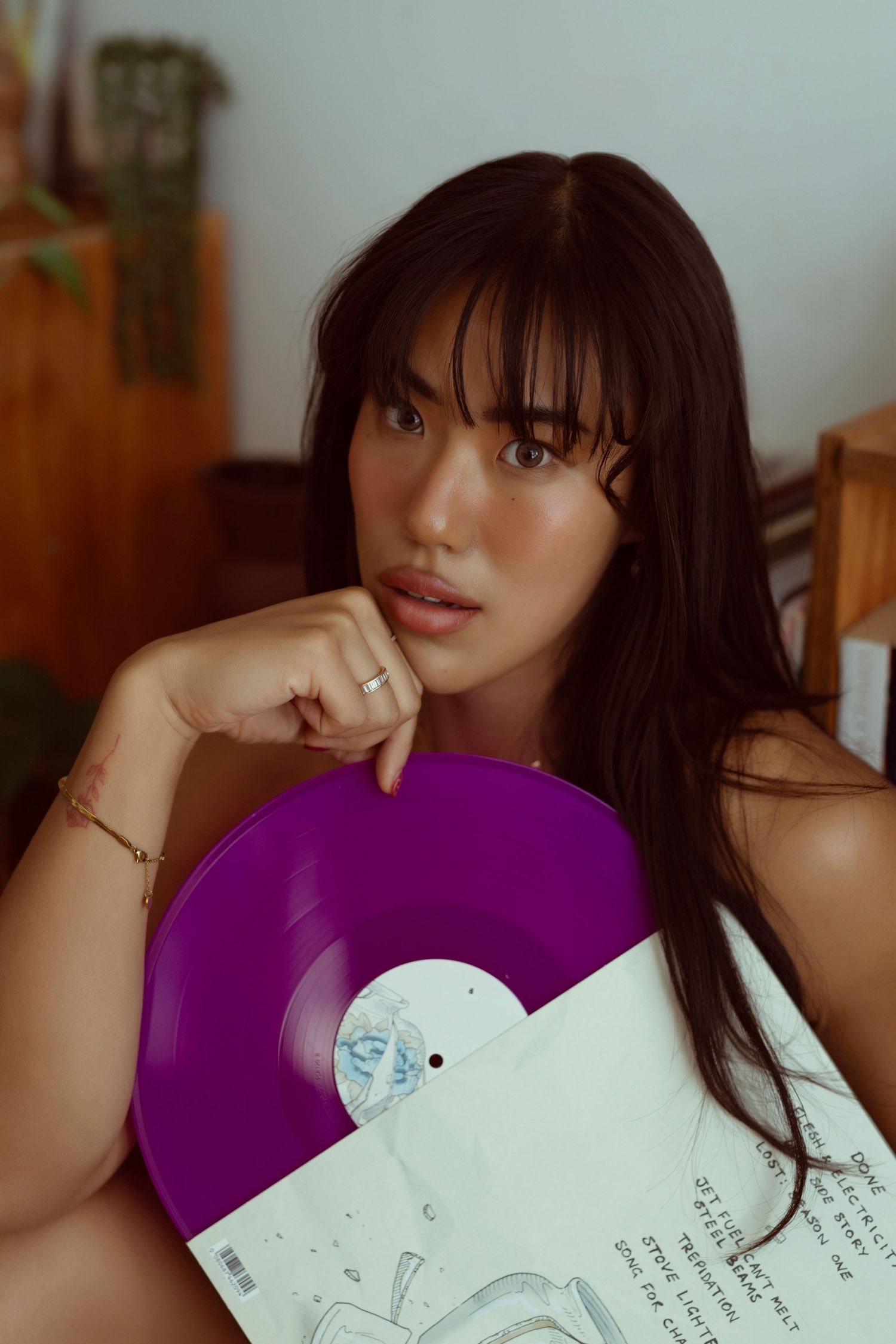 Young woman with long dark hair holding a purple vinyl record album, wearing gold jewelry, in a cozy room.