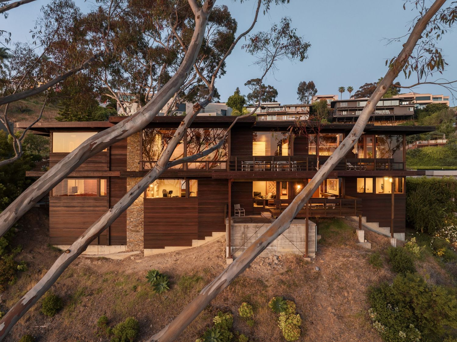Eucalyptus trees frame the tiered rear facade of 7226 Romero in La Jolla, a midcentury Liebhardt home with glowing decks at dusk.