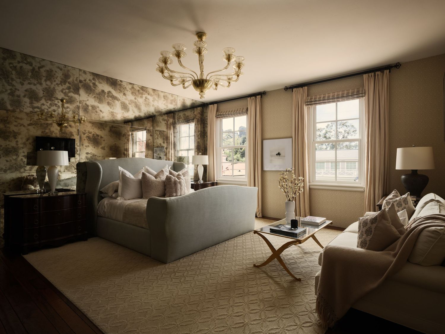 Primary bedroom suite at 7757 Whitefield with upholstered bed, sitting area, mirrored accent wall, and soft daylight through tall windows.