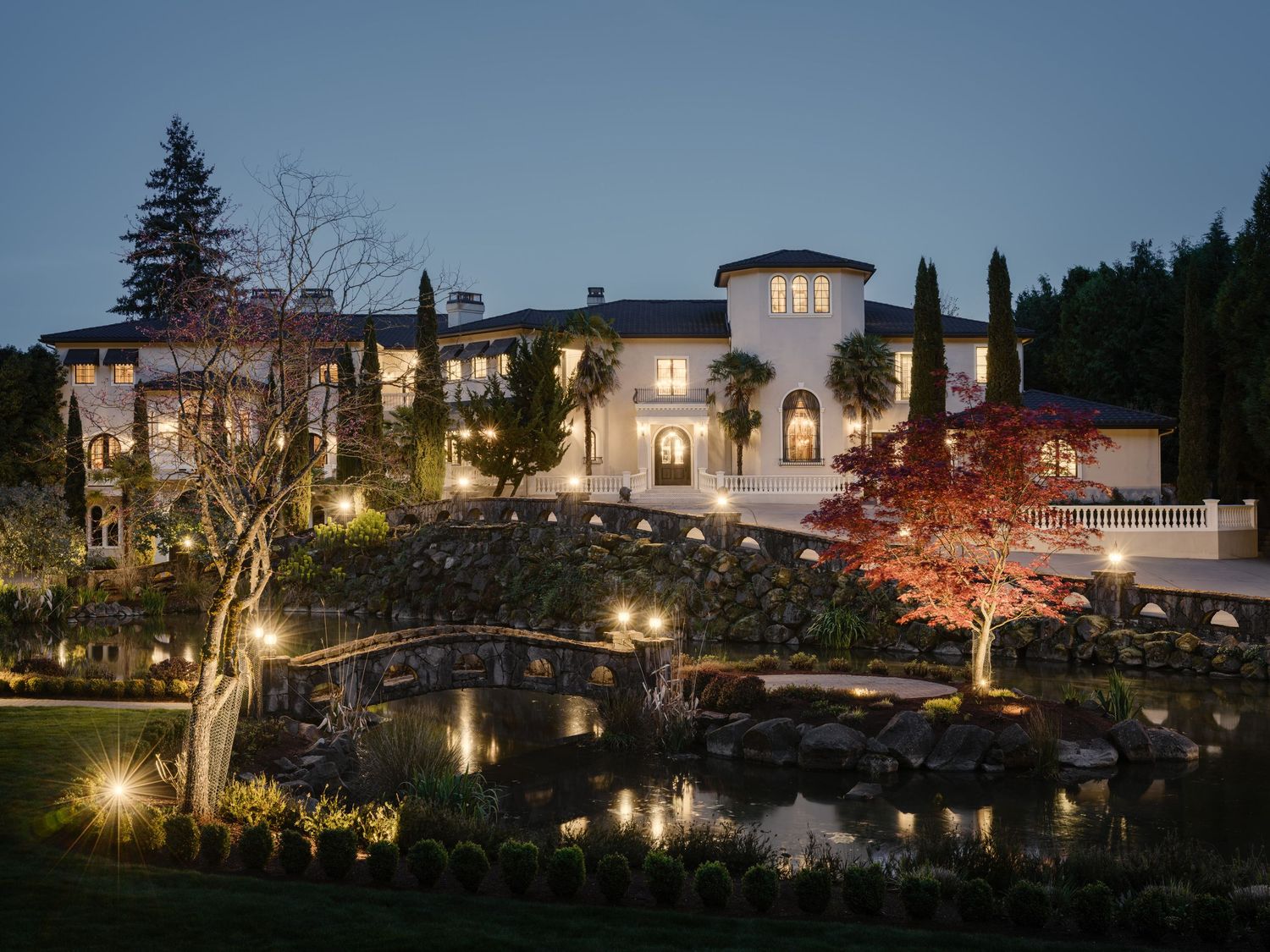 Night exterior of Fielding House with illuminated landscaping, stone bridge, and reflective pond creating an atmospheric luxury scene.