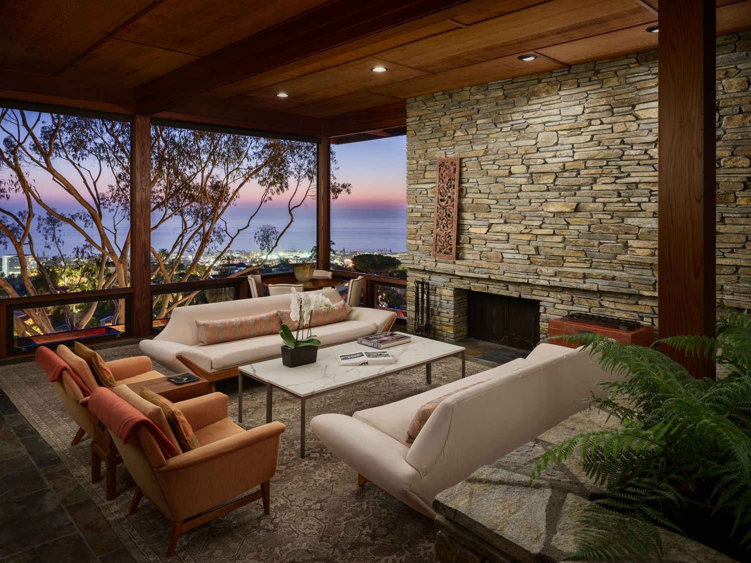 Ocean view living room at 7226 Romero, with wood ceilings, stone fireplace, modern seating, and eucalyptus framing the La Jolla coastline.