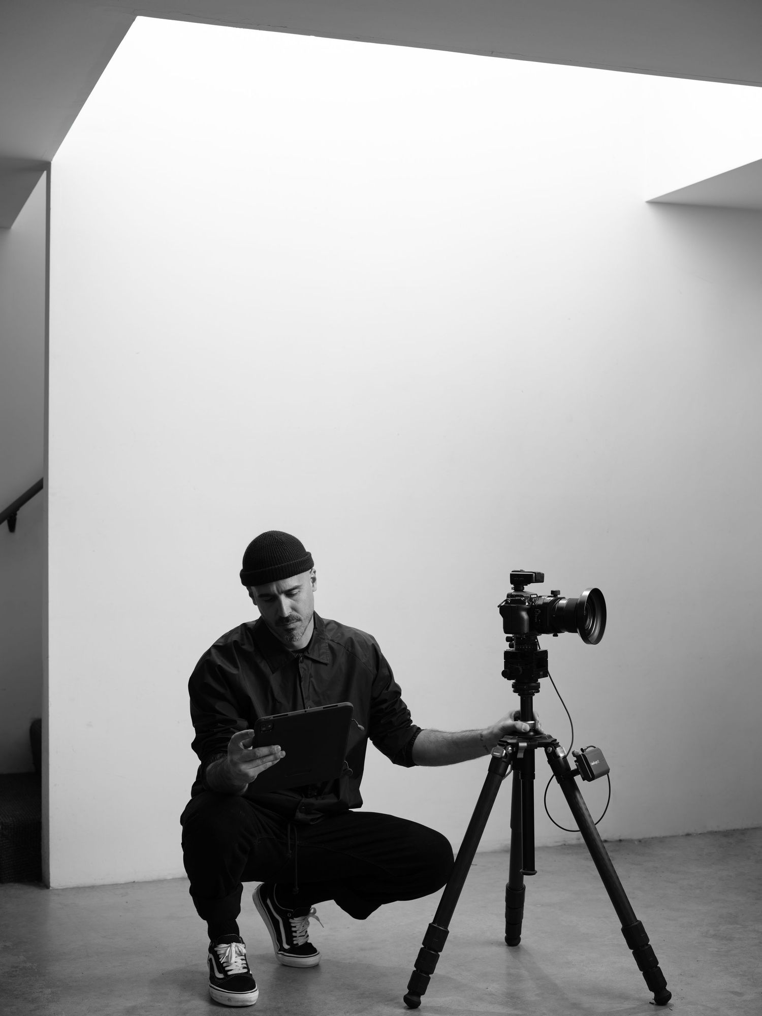 Luca Prodi seated with camera and tripod in a minimalist architectural space, captured in black and white.