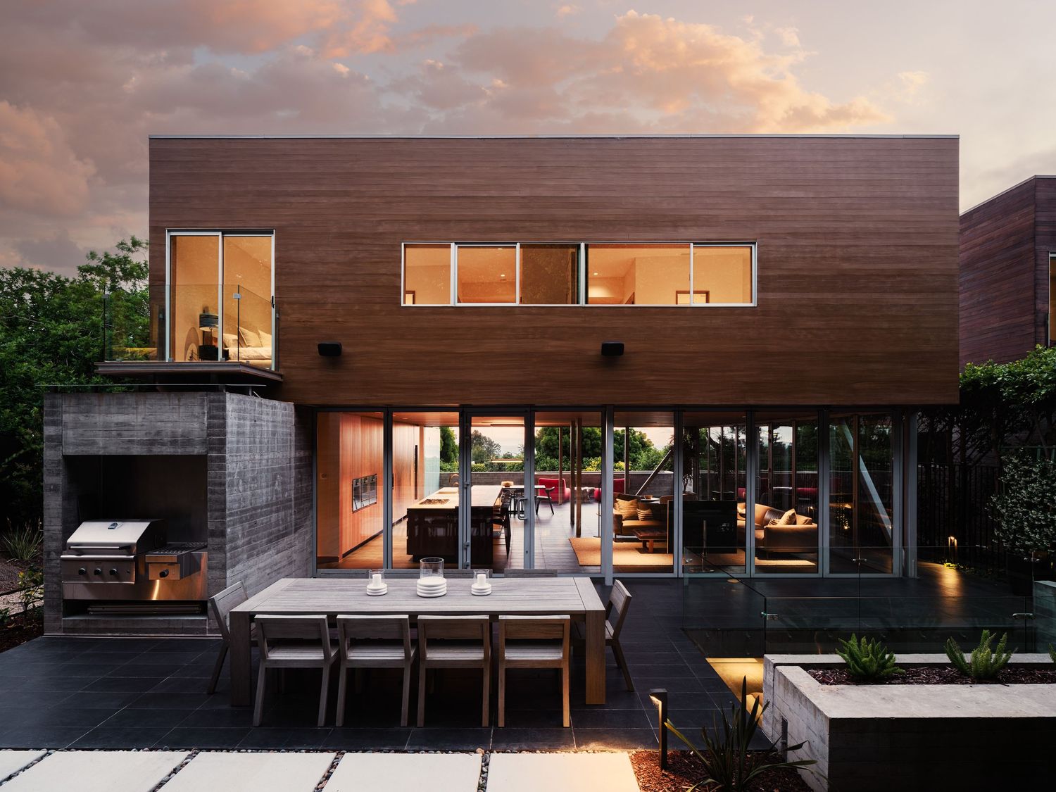 Rear exterior of 7003 Fay Avenue with floor-to-ceiling glass, indoor-outdoor living, and warm modern architecture at dusk.