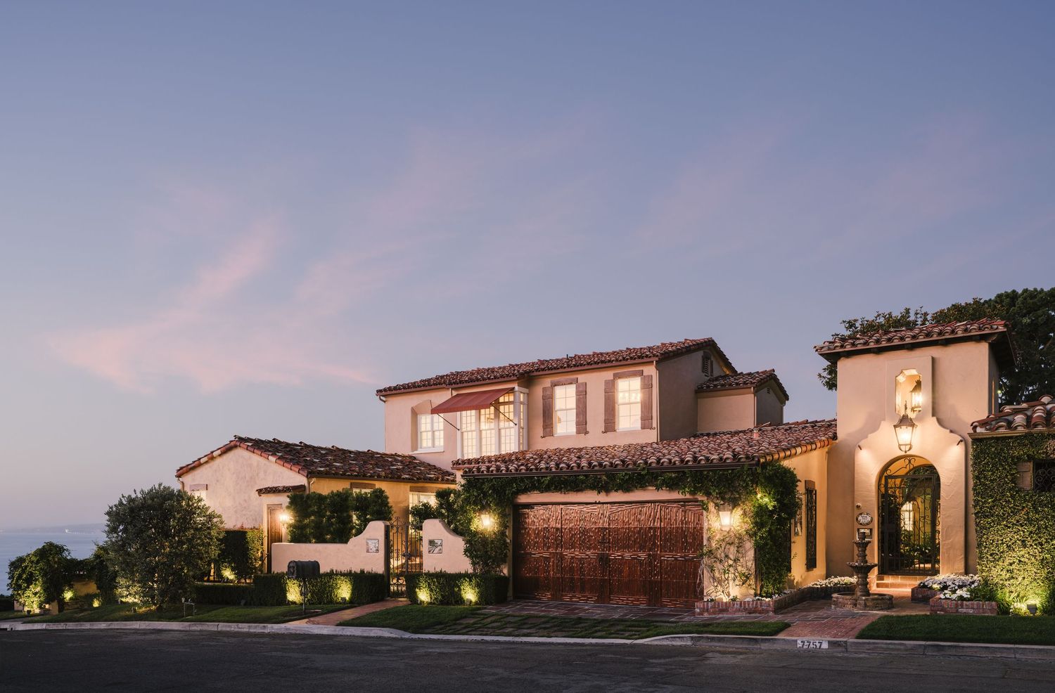 Twilight exterior of a luxury Mediterranean La Jolla residence with ocean views, clay tile roof, landscaped entry, and warm architectural li