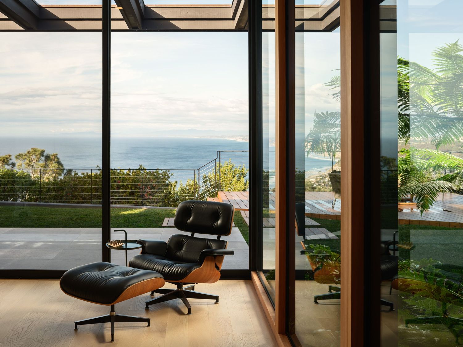 Interior lounge at 7404 Hillside Drive framed by floor-to-ceiling glass, opening to landscaped grounds and distant coastal views.