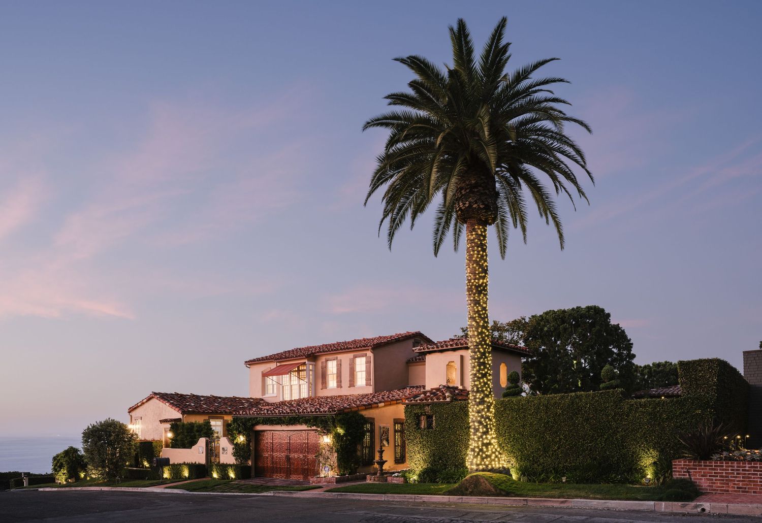 Twilight exterior of a luxury Mediterranean-style La Jolla home with ocean views, palm tree, manicured hedges, and architectural lighting.