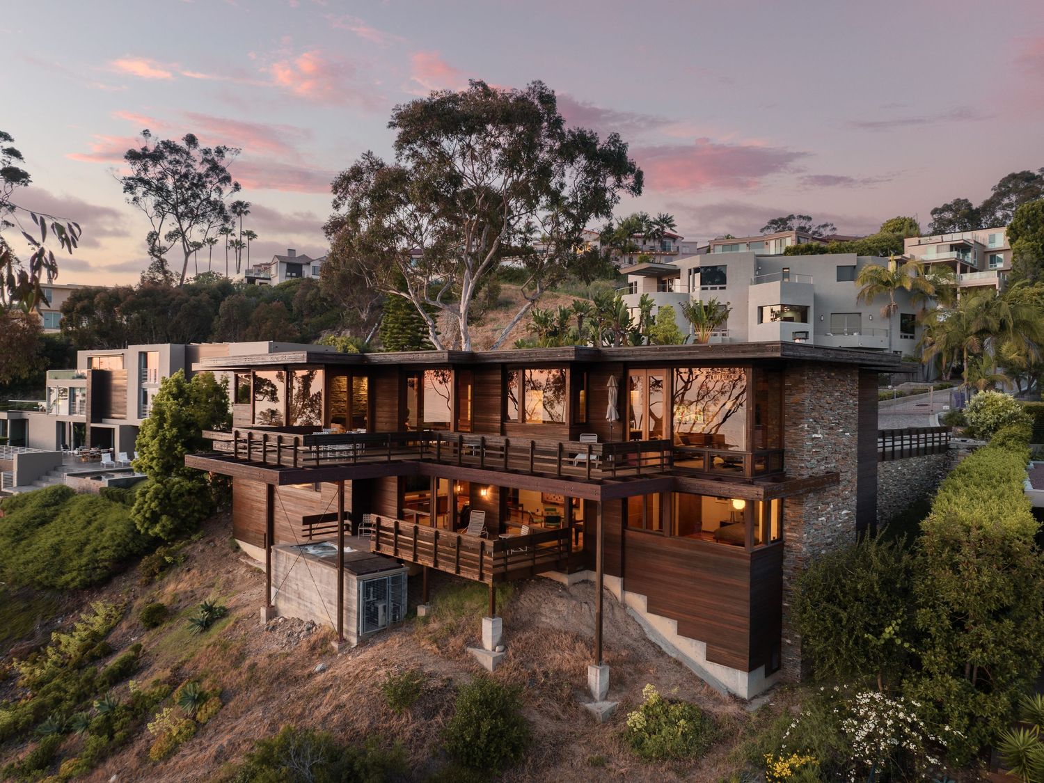 Sunset rear view of 7226 Romero midcentury home in La Jolla, showing cantilevered decks, warm wood cladding, and steep hillside site.