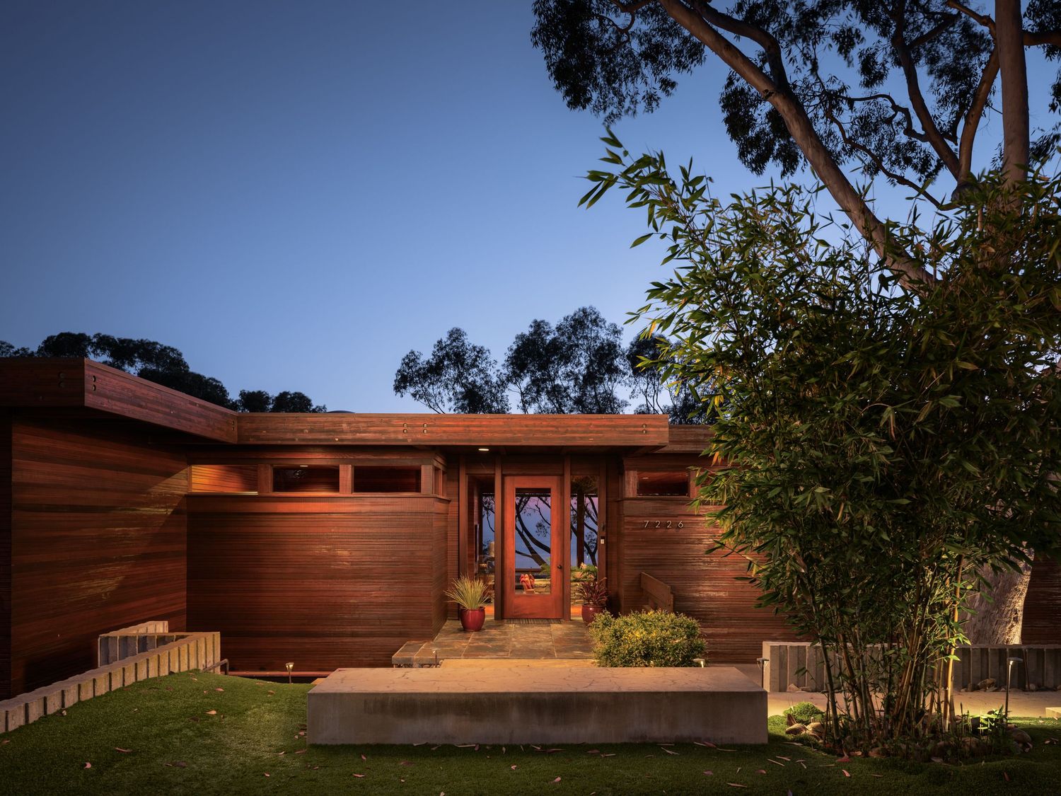 Dusk exterior of 7226 Romero, a midcentury La Jolla luxury residence with cedar siding, uplighting, and mature eucalyptus at the entry.