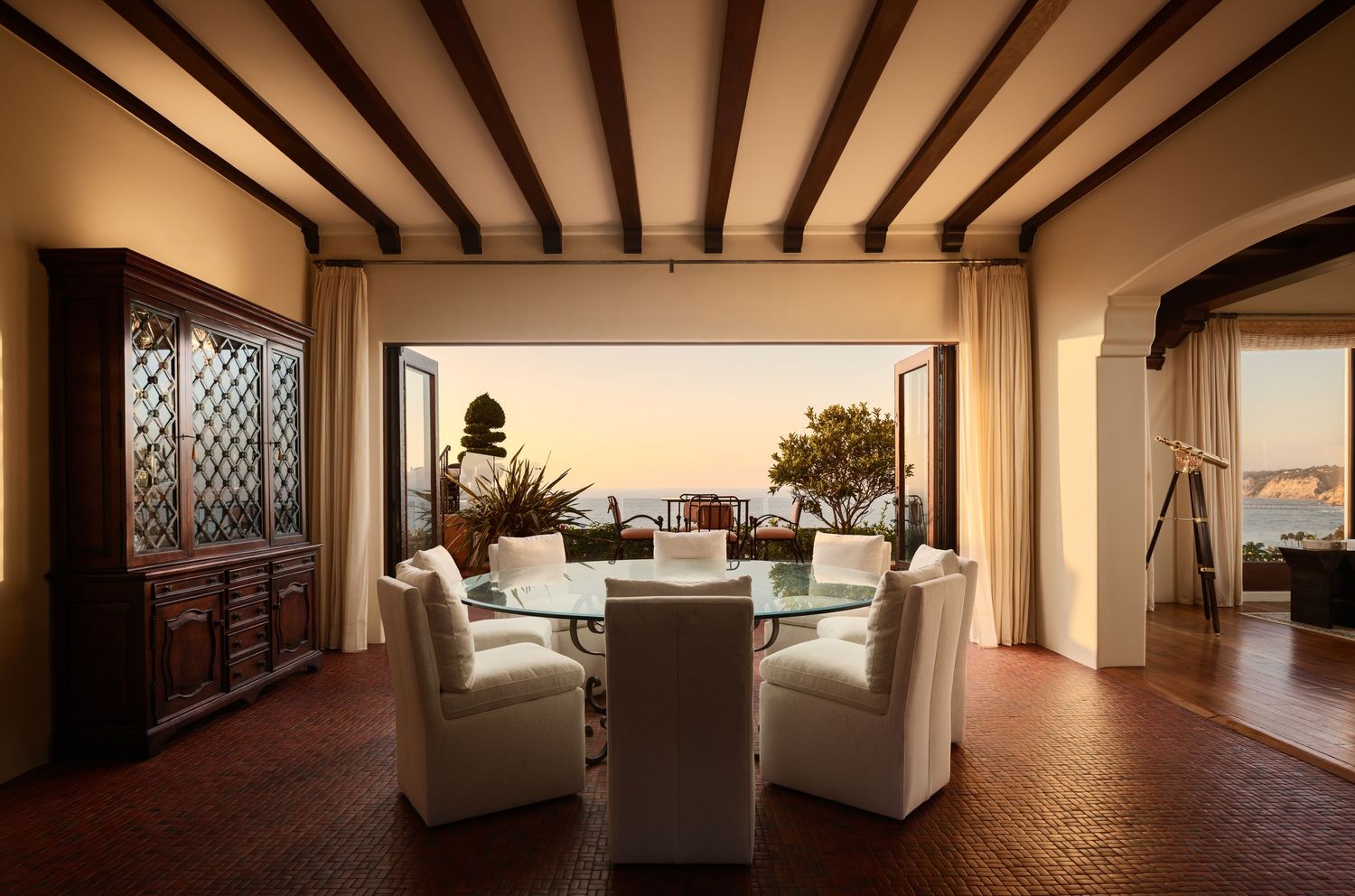 Luxury real estate photo of Casa De La Vista dining area with open French doors revealing ocean views, wood beams, and golden sunset light.